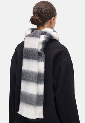 Écharpe en laine avec des rayures alternées en gris et blanc, dotée de bords frangés. Portée sur un manteau noir, mettant en valeur une finition douce et texturée.