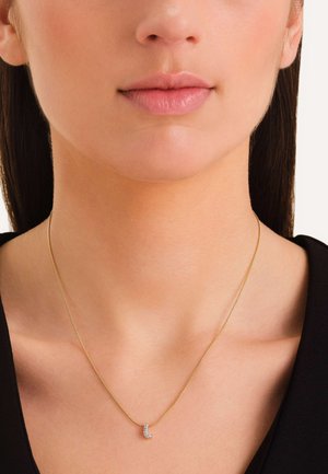 Gouden ketting met een delicate ketting en een kleine, gebogen hanger met heldere edelstenen, gedragen tegen een donkere shirt.