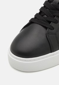 Sneaker en cuir synthétique noir avec un dessus lisse, bout rond, semelle en caoutchouc blanc texturé et lacets noirs.