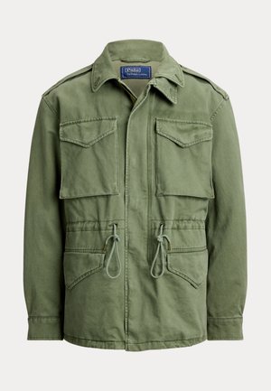 Joven con chaqueta verde, camisa azul, pantalones beige de cuadros, zapatillas blancas, sosteniendo un gran bolso tote de cuero marrón, de pie frente a un fondo neutro.