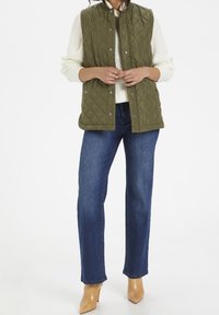 Persona con chaleco acolchado color verde oliva sobre suéter blanco, jeans rectos azules y botas de tacón de punta en color beige sobre fondo blanco.