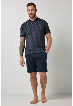 Mand med tatoveret arm iført navy mønstret kortærmet skjorte og navy shorts, stående barfodet mod en ensfarvet baggrund.