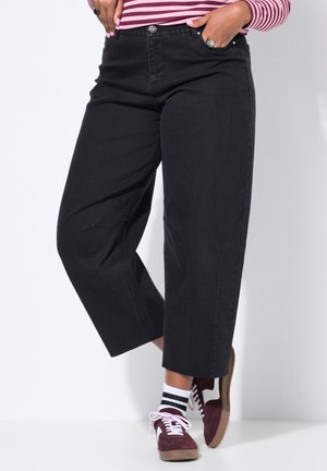 Schwarze weit geschnittene Hosen mit leichter Textur, seitlichen Taschen und einem Knopfverschluss, kombiniert mit burgunderfarbenen Sneakers und gestreiften Socken.