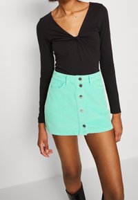 Bodysuit negro de manga larga con escote en V y detalle de nudo, combinado con una falda de pana verde menta que cuenta con un cierre de botones en la parte delantera y bolsillos.