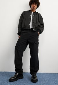 Μαύρο μπουφάν bomber με κουμπιά snap, συνδυασμένο με μαύρο παντελόνι cargo με τσέπες. Τα μαύρα παπούτσια με χοντρή σόλα ολοκληρώνουν την εμφάνιση.