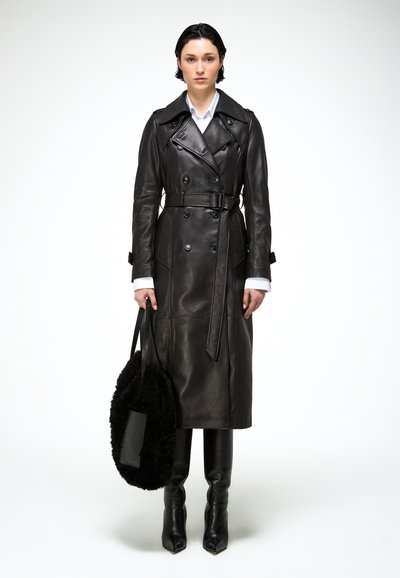 Femme debout portant un long trench-coat en cuir noir ceinturé, des bottes noires à bout pointu, tenant un grand sac fourre-tout noir et duveteux.