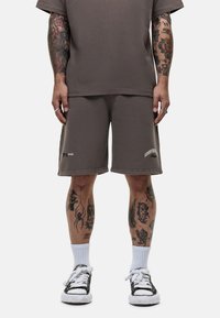 T-shirt in cotone marrone e pantaloni corti abbinati. I pantaloni corti presentano del testo stampato sul lato. Calzini bianchi alla caviglia e sneakers nere completano il look.