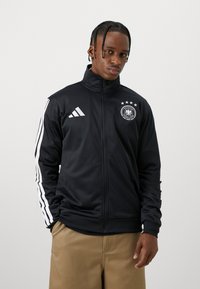 Μαύρο αθλητικό μπουφάν με ψηλό γιακά, με λευκές κατακόρυφες ρίγες και λογότυπο της γερμανικής εθνικής ομάδας στο στήθος. Λείο, ελαφρύ ύφασμα.