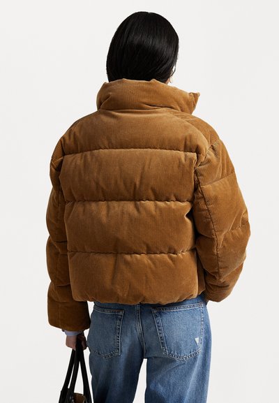 Jachetă puffer din mătase corduroy maro, cu guler înalt, având secțiuni quilted. Purtați cu blugi albaștri și ținând o geantă de mână neagră.