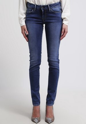 Jeans ajustados en denim azul oscuro, con bolsillos frontales, cremallera y un acabado ligeramente desgastado. Combinados con zapatos de tacón de estampado.