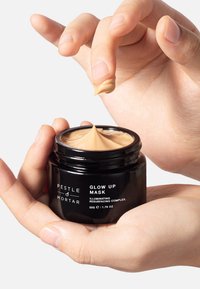 Schwarzes Glas mit "Glow Up Mask" und einer beige-cremigen Textur, gehalten von einer Hand. Das Etikett enthält den Produktnamen und eine Beschreibung.