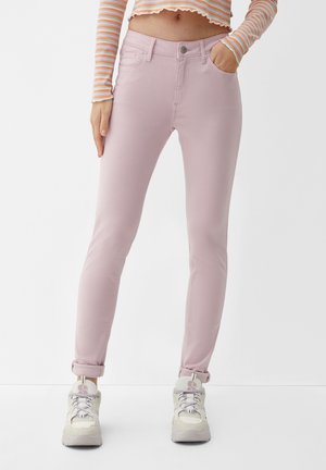 Schmal geschnittene, hellrosafarbene Hose mit umgeschlagenen Beinabschlüssen, kombiniert mit weißen Turnschuhen und einem kurz geschnittenen, gestreiften Langarmoberteil.