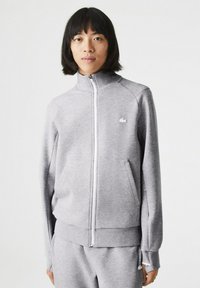 Chaqueta gris con cremallera en tejido suave y alto collar. Presenta bolsillos laterales, puños acanalados y un pequeño logo blanco en el pecho.