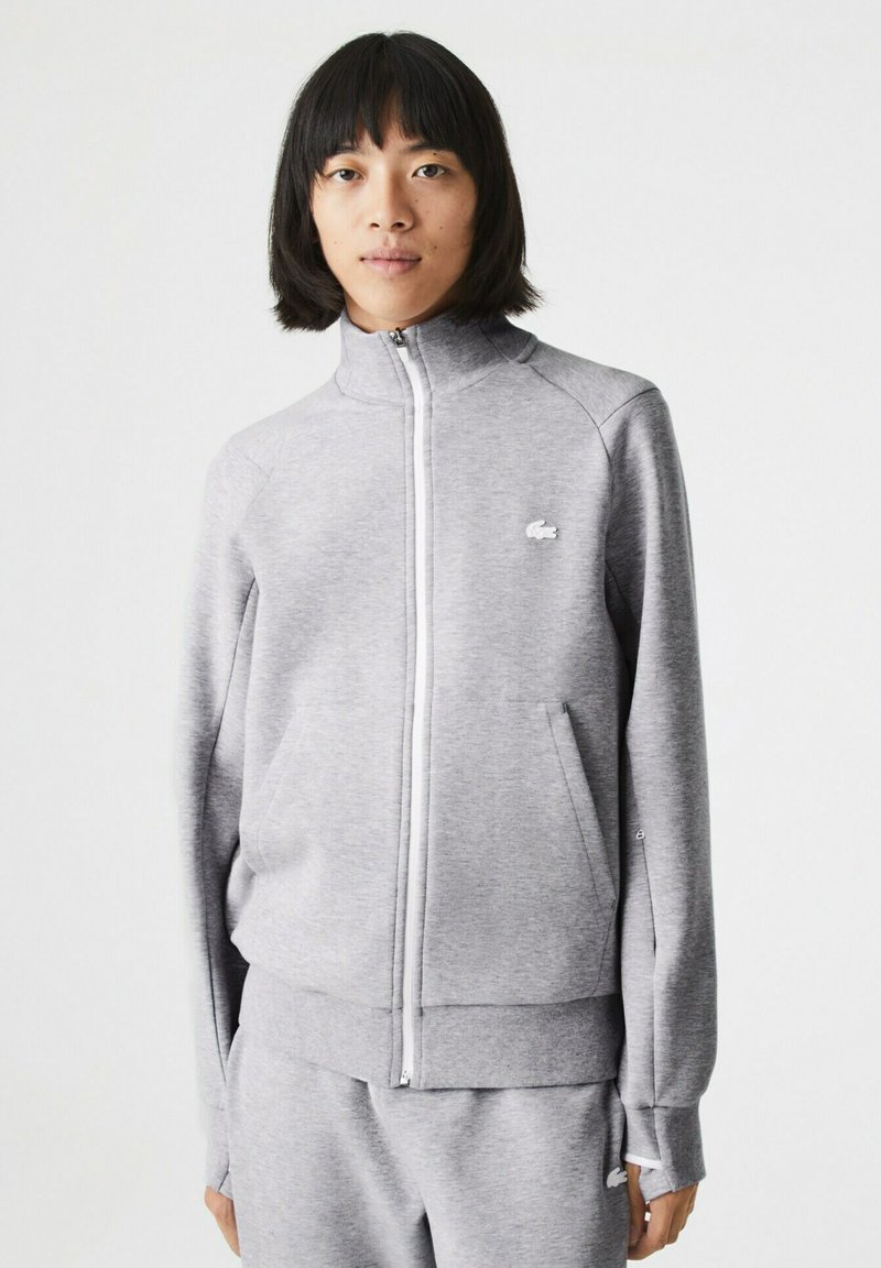 Chaqueta gris con cremallera en tejido suave y alto collar. Presenta bolsillos laterales, puños acanalados y un pequeño logo blanco en el pecho.