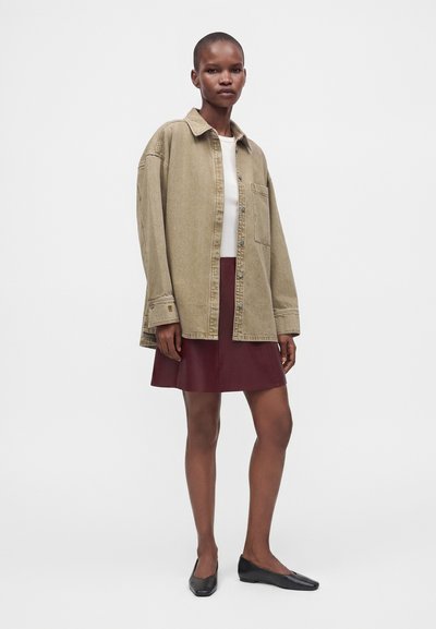 Άτομο που φοράει ένα oversized μπεζ πουκάμισο με κουμπιά πάνω από ένα λευκό τοπ, βουργουνδί φούστα και μαύρα επίπεδα παπούτσια, στέκεται μπροστά σε απλό φωτεινό φόντο.