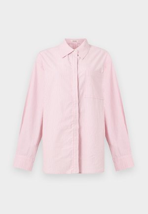 Femme debout les mains dans les poches, portant une chemise ample rayée rose, un short crème et des sandales beiges avec des détails en nœud.