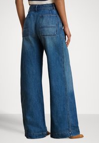 Vide, blå denimjeans med middels høy midje, med to baklommer og en falmet finish. Stoffet har en glatt tekstur.