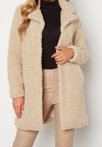Beige fluffy frakke med klapkrave, lange ærmer og forreste knapper, lavet af tekstureret stof, båret over en sort top og beige bukser.
