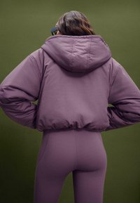 Kürzende Daunenjacke in Mauve mit Kapuze, ausgestattet mit einer glatten Stofftextur und gerippten Säumen, kombiniert mit passenden enganliegenden Leggings.
