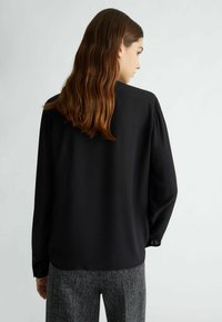 Blusa nera con una texture liscia, dotata di maniche lunghe e polsini abbottonati. Il retro presenta un design ordinato e su misura. Abbinata a pantaloni grigi con motivo.
