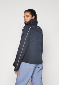 Veste matelassée marine avec col haut, accents de passepoil blanc et patch logo sur l'épaule. Tissu lisse avec texture matelassée.