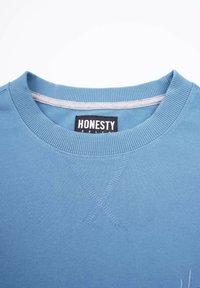Blå bomullströja med ribbad rund halsringning, tvärstygnsdetaljer och en svart etikett med "HONESTY" vid halsringningen.