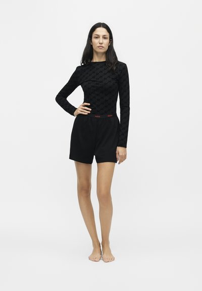Femme debout pieds nus, cheveux longs et foncés, portant un haut noir à manches longues et un short noir avec une ceinture rouge "Hugo".