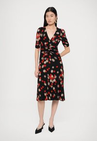 Musta mekko, jossa on lyhyet hihat ja kuvana nainen. Kangas on värikkäällä, pilkullisella kuviolla, jossa on punaista ja beigeä, ja siinä on V-pääntie.