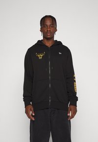 Sudadera con cremallera en negro, con un logo de toro en amarillo en el pecho, que cuenta con un bolsillo frontal y texto en la manga derecha, confeccionada en una tela suave.