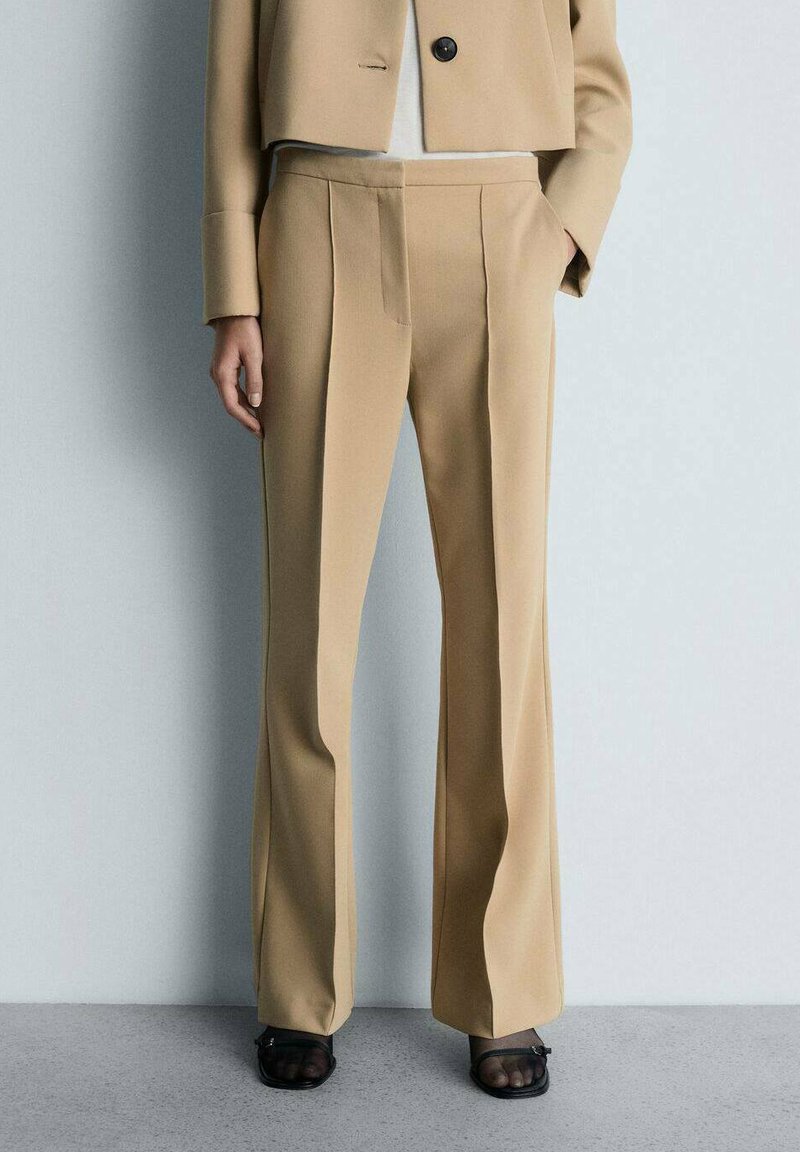 Pantaloni beige su misura con davanti piatto, tasche laterali e gamba leggermente svasata; il tessuto ha una texture liscia. Abbinati a una giacca corta.