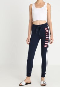 Pantalones de chándal azul marino con franjas laterales blancas y texto rojo de "Hollister". Combinados con una camiseta sin mangas corta blanca y sandalias tipo slide.