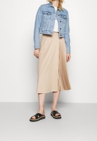 Chaqueta de mezclilla en azul claro, estilo cropped. Camiseta blanca por debajo. Falda midi plisada en color beige. Sandalias negras con tiras anchas.