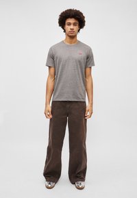 Camiseta de algodón gris con mangas cortas y un pequeño logo rojo, combinada con pantalones anchos marrones y zapatos metálicos. Fondo liso.