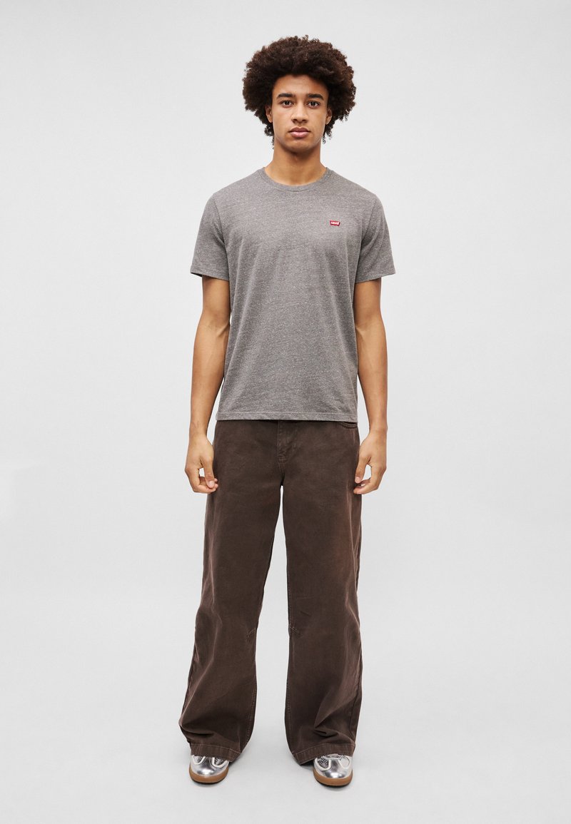 Szara bawełniana koszulka z krótkim rękawem i małym czerwonym logo, w połączeniu z brązowymi spodniami o szerokim kroju i metalicznymi butami. Gładkie tło.