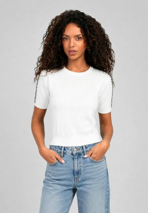 Femme aux cheveux bouclés portant un haut blanc à manches courtes avec des coutures noires sur les manches et un jean bleu taille haute, mains dans les poches.