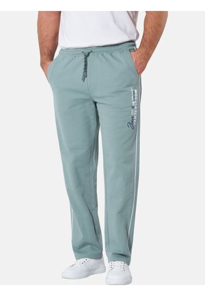 Mann trägt hellgrüne Jogginghose mit Seitentaschen und weißen Sneakers, steht mit den Händen in den Taschen vor neutralem Hintergrund.