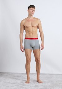 Sivé boxerky s červeným pásom Nike, ktoré majú tesný strih a hladkú textúru, vystavené na stojacom mužskom modeli na jednofarebnom pozadí.