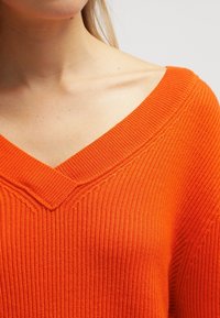V-Ausschnittpullover in leuchtendem Orange, mit gerippter Textur und lockerer Passform, die den Ausschnitt und die Schultern betont.
