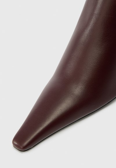 Zapato de tacón alto de cuero burdeos con punta afilada y textura suave, que presenta un diseño elegante. El borde inferior es ligeramente redondeado.