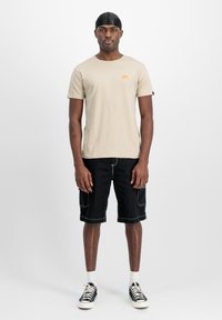 Beige Baumwoll-T-Shirt mit orangefarbenem Logo, kombiniert mit schwarzen Cargo-Shorts mit Seiten Taschen und weißen Nähten, getragen mit schwarzen Sneakers.
