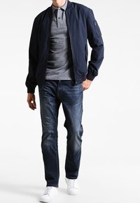 Marinkappsjacka med ribbad krage, fickor med dragkedja, grå polotröja under, mörk denimjeans och vita sneakers. Avslappnad outfit.