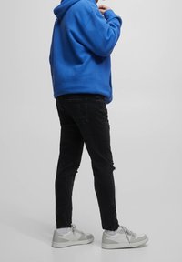 Osoba v modré mikině s kapucí, černých roztrhaných džínách, bílých ponožkách a bílých teniskách stojící na jednobarevném světle šedém pozadí.