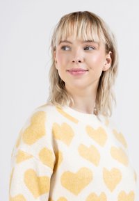 Maglione con una base crema e motivi a cuore gialli, realizzato in materiale lavorato a maglia, con scollatura rotonda e vestibilità comoda.