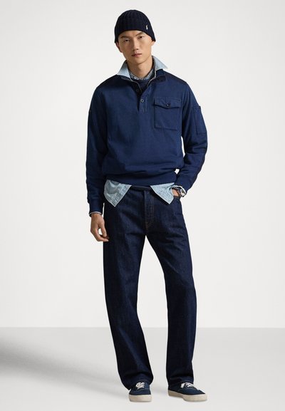 Temno modra majica z dolgimi rokavi, ovratnikom in žepom na prsih, oblečena preko svetlo modre denim srajce, kombinirana s temnimi kavbojkami in temno modro obutvijo.