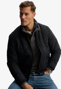 Homme portant une veste matelassée noire sur un pull gris et une chemise à carreaux, jean bleu, une main dans la poche et regardant sur le côté.