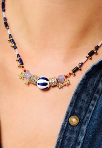 Collier perlé composé de pierres bleu, doré et violet, avec une perle centrale blanche ornée de rayures bleues et d'accents floraux.