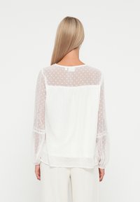 Blouse semi-transparente blanche avec un motif à pois, manches longues bouffantes et détails en dentelle aux poignets. La vue de dos montre un col rond.