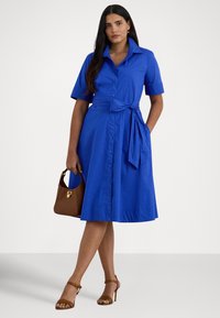 Ženska stoji v modri obleki s gumbi in pasom v pasu, drži rjavo torbico in nosi rjave sandale z visoko peto na belem ozadju.