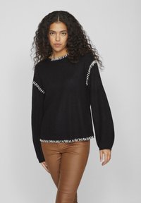 Čierny pletený sveter s voľným strihom, ktorý obsahuje biele akcentové prešívanie pozdĺž švov a rebrovanú textúru. Dlhé rukávy a široký výstrih.