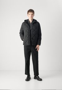Jaunas vyras, apsirengęs juoda siuvinėta striuke, juodu zip-up marškinėliais, juodomis tiesiomis kelnėmis ir juodais odiniais batais, stovi prieš baltą foną.